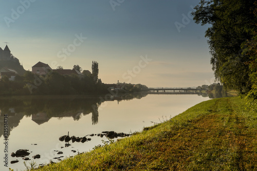 River bank of Drava at the city of Ptuj on an early summer morning. Visible reflection on the water and bridges in the distance. Nobody on the green grass shore. photo