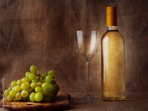 Claster of fresh juicy white grapes on a wooden board and glass and bottle of white wine on hessian cloth background. Still life, Wine making concept.