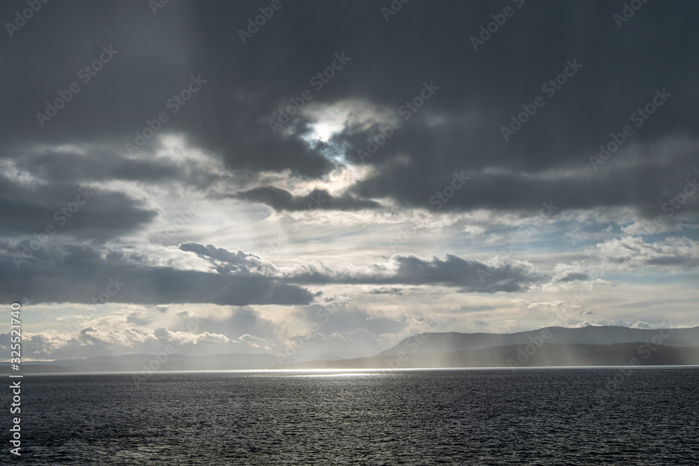 sunlight piercing through heavy cloud over the horizon illuminating the water surface on the ocean with mist of water surrounding the nearby island