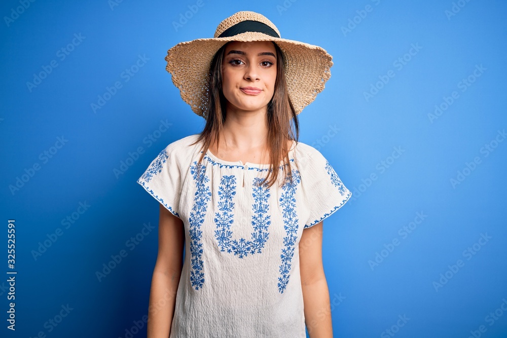 Young beautiful brunette woman wearing casual t-shirt and summer hat over blue background with serious expression on face. Simple and natural looking at the camera.