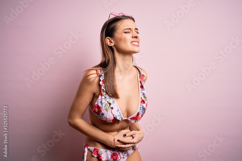 Young beautiful woman on vacation wearing bikini and sunglasses over pink background with hand on stomach because nausea, painful disease feeling unwell. Ache concept.