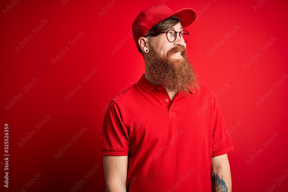 Young handsome delivery man wearing glasses and red cap over isolated background looking away to side with smile on face, natural expression. Laughing confident. Young handsome delivery man wearing glasses and red cap over isolated background looking away to side with smile on face, natural expression. Laughing confident.