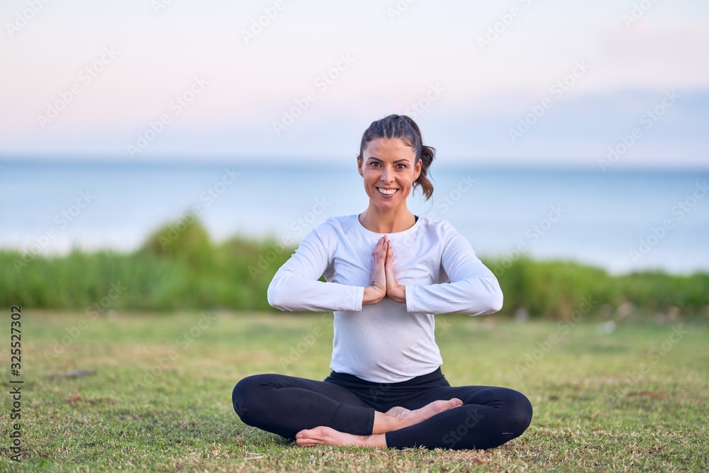 Young beautiful sportwoman smiling happy practicing yoga. Coach sitting with smile on face teaching prayer pose at park