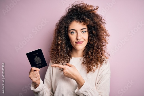 Beautiful tourist woman with curly hair and piercing holding canada canadian passport id very happy pointing with hand and finger