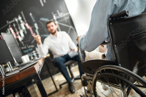 Cropped photo of office worker in a wheelchair discussing something with his male colleague while working together in the modern office
