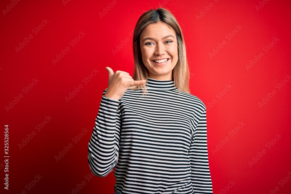 Young beautiful blonde woman wearing casual striped sweater over red isolated background smiling doing phone gesture with hand and fingers like talking on the telephone. Communicating concepts. Young beautiful blonde woman wearing casual striped sweater over red isolated background smiling doing phone gesture with hand and fingers like talking on the telephone. Communicating concepts.