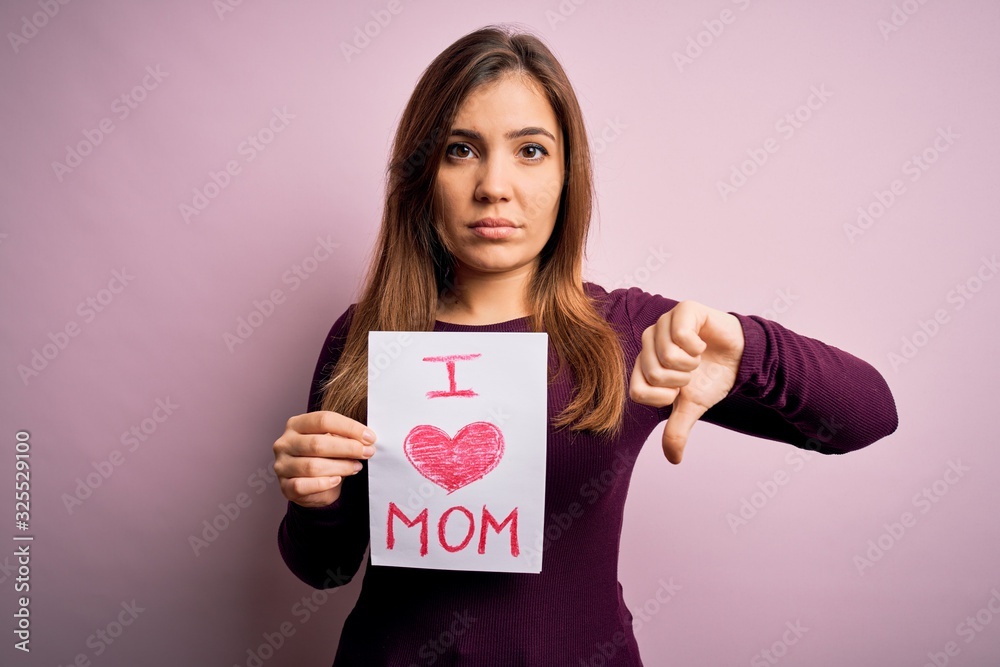 Young beautiful woman holding paper with love mom message celebrating mothers day with angry face, negative sign showing dislike with thumbs down, rejection concept Young beautiful woman holding paper with love mom message celebrating mothers day with angry face, negative sign showing dislike with thumbs down, rejection concept