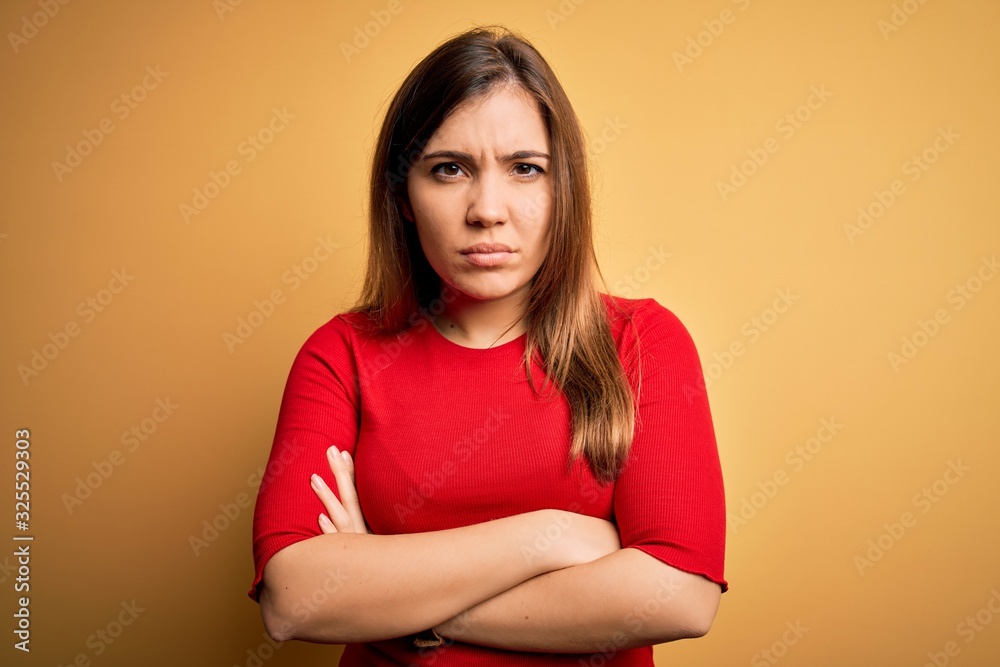 Beautiful young woman wearing casual red t-shirt over yellow isolated background skeptic and nervous, disapproving expression on face with crossed arms. Negative person.
