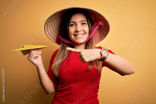 Tourist woman wearing traditional asian rice paddy straw hat holding paper plane for a trip very happy pointing with hand and finger