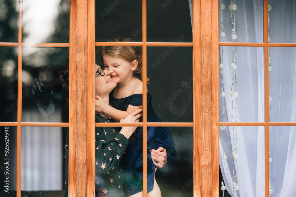 Handsome woman with pretty face hugs a beautiful daughter and looks through big window Handsome woman with pretty face hugs a beautiful daughter and looks through big window