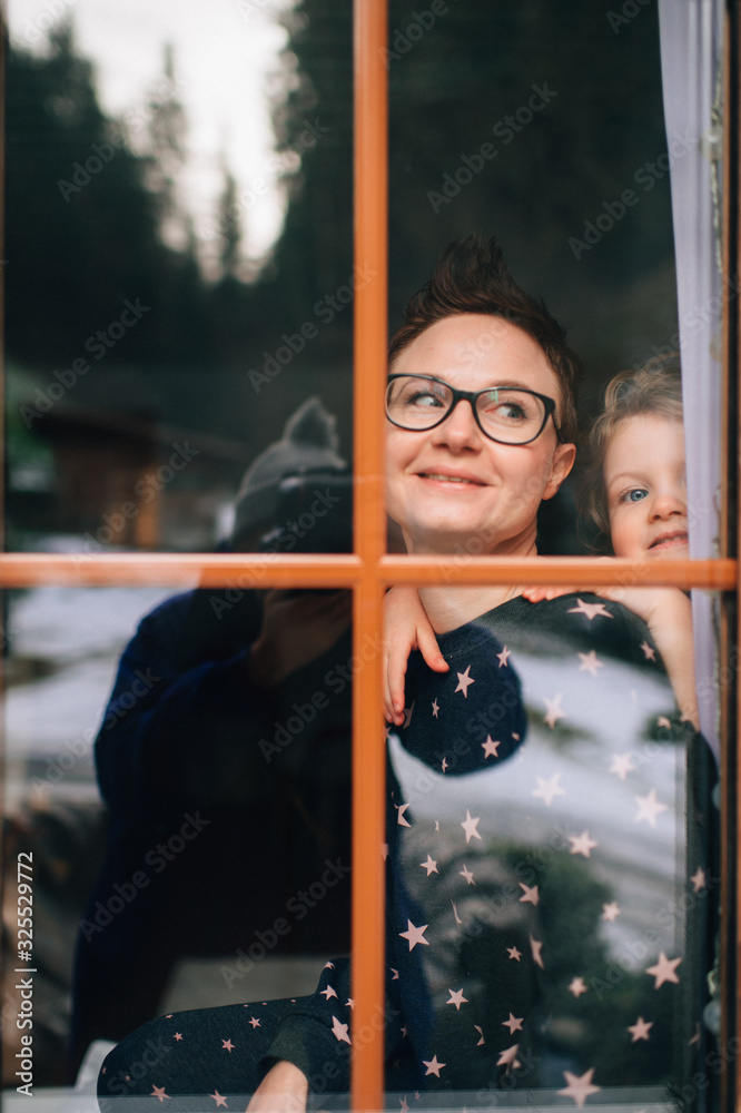 Handsome woman with pretty face hugs a beautiful daughter and looks through big window