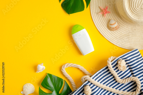 Moisturizer sunscreen lotion bottle without labels, striped beach bag, tropical leaves, female hat and seashells on yellow background. Summer skin care cosmetic product and sun protection concept