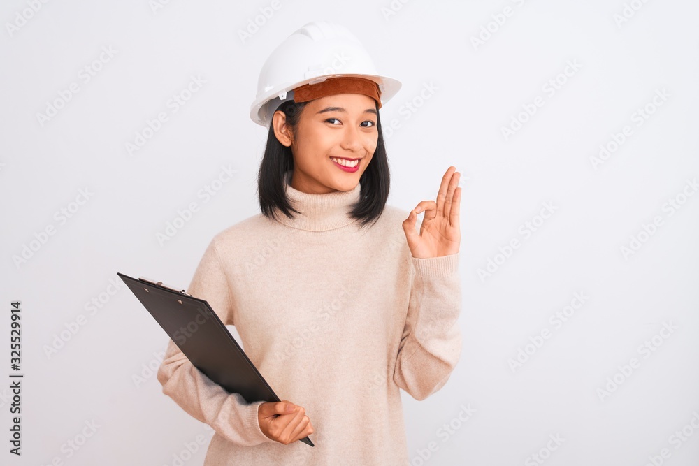 Chinese architect woman wearing helmet holding clipboard over isolated white background doing ok sign with fingers, excellent symbol Chinese architect woman wearing helmet holding clipboard over isolated white background doing ok sign with fingers, excellent symbol