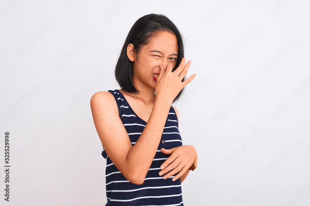 Young chinese woman wearing striped t-shirt standing over isolated white background smelling something stinky and disgusting, intolerable smell, holding breath with fingers on nose. Bad smells concept Young chinese woman wearing striped t-shirt standing over isolated white background smelling something stinky and disgusting, intolerable smell, holding breath with fingers on nose. Bad smells concept