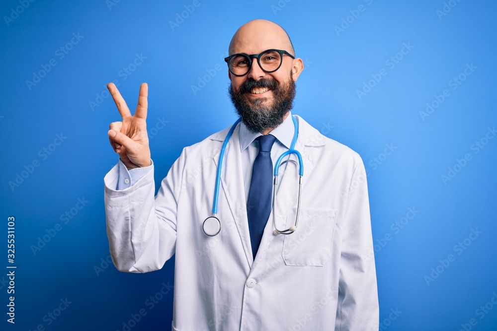 Handsome bald doctor man with beard wearing glasses and stethoscope over blue background smiling with happy face winking at the camera doing victory sign with fingers. Number two.