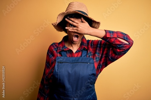 Young African American afro farmer woman with curly hair wearing apron and hat peeking in shock covering face and eyes with hand, looking through fingers with embarrassed expression.
