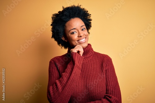 Young beautiful African American afro woman with curly hair wearing casual turtleneck sweater looking confident at the camera smiling with crossed arms and hand raised on chin. Thinking positive. © Krakenimages.com