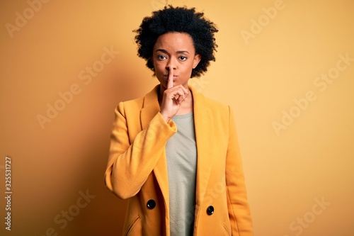 Young beautiful African American afro businesswoman with curly hair wearing yellow jacket asking to be quiet with finger on lips. Silence and secret concept. © Krakenimages.com