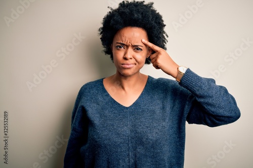 Young beautiful African American afro woman with curly hair wearing casual sweater pointing unhappy to pimple on forehead, ugly infection of blackhead. Acne and skin problem
