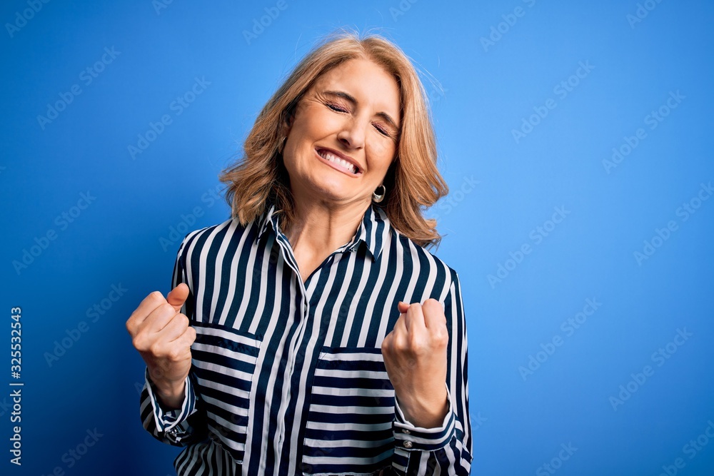 Middle age beautiful blonde woman wearing casual striped shirt standing over blue background very happy and excited doing winner gesture with arms raised, smiling and screaming for success. Middle age beautiful blonde woman wearing casual striped shirt standing over blue background very happy and excited doing winner gesture with arms raised, smiling and screaming for success.
