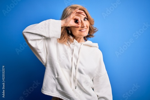 Middle age beautiful blonde sportswoman wearing sportswear over isolated blue background doing ok gesture with hand smiling, eye looking through fingers with happy face.