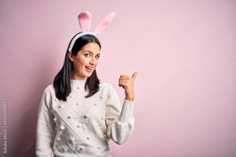 Young caucasian woman wearing cute easter rabbit ears over pink isolated background smiling with happy face looking and pointing to the side with thumb up. Young caucasian woman wearing cute easter rabbit ears over pink isolated background smiling with happy face looking and pointing to the side with thumb up.