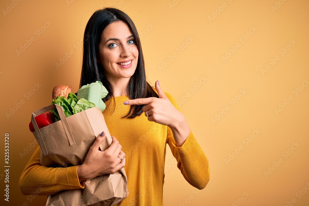 Young brunette woman with blue eyes holding healthy groceries in paper bag from supermarket very happy pointing with hand and finger Young brunette woman with blue eyes holding healthy groceries in paper bag from supermarket very happy pointing with hand and finger