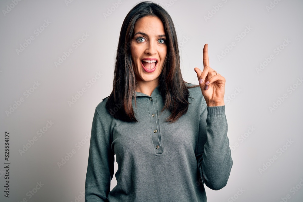 Young brunette woman with blue eyes wearing casual green sweater over white background pointing finger up with successful idea. Exited and happy. Number one.