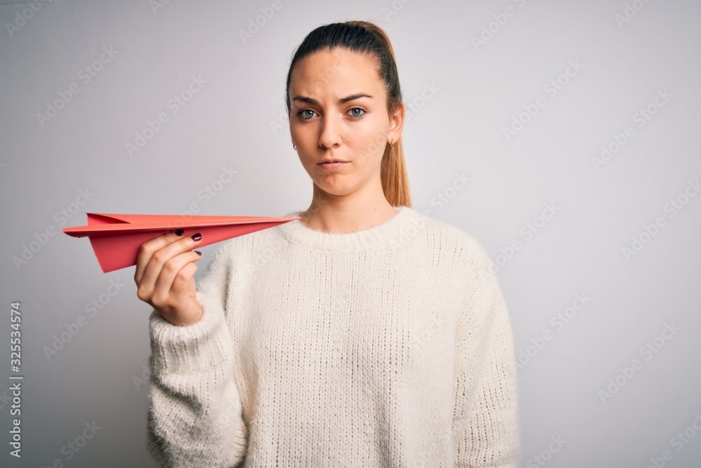 Beautiful blonde woman with blue eyes holding red paper airplane over white background with a confident expression on smart face thinking serious Beautiful blonde woman with blue eyes holding red paper airplane over white background with a confident expression on smart face thinking serious