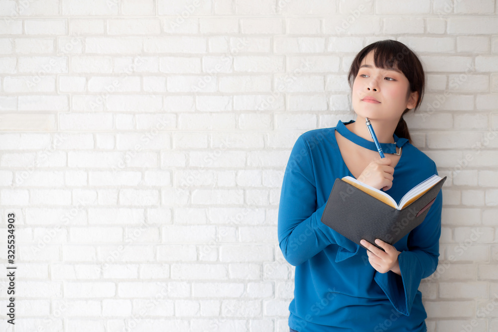 Beautiful asian woman smiling standing thinking and writing notebook on concrete cement white background at home, girl homework on book, education and lifestyle concept. Beautiful asian woman smiling standing thinking and writing notebook on concrete cement white background at home, girl homework on book, education and lifestyle concept.