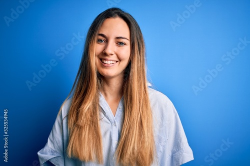 Young beautiful blonde woman with blue eyes wearing striped shirt over blue background with a happy and cool smile on face. Lucky person.