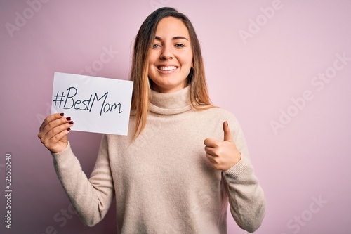 Young beautiful woman holding paper with best mom message celebrating mothers day happy with big smile doing ok sign, thumb up with fingers, excellent sign