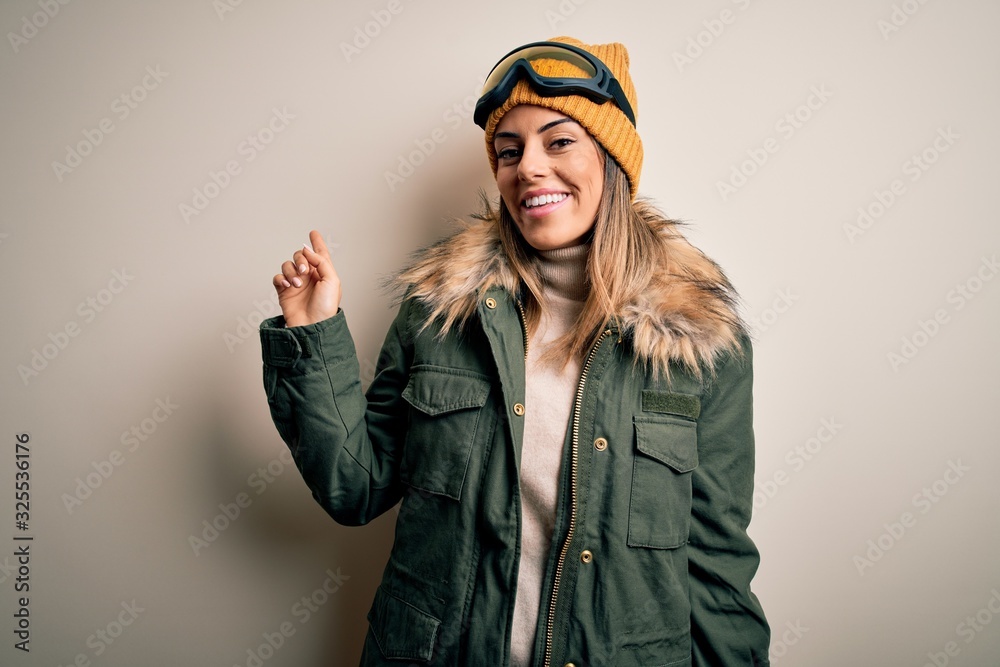 Young brunette skier woman wearing snow clothes and ski goggles over white background with a big smile on face, pointing with hand finger to the side looking at the camera. Young brunette skier woman wearing snow clothes and ski goggles over white background with a big smile on face, pointing with hand finger to the side looking at the camera.