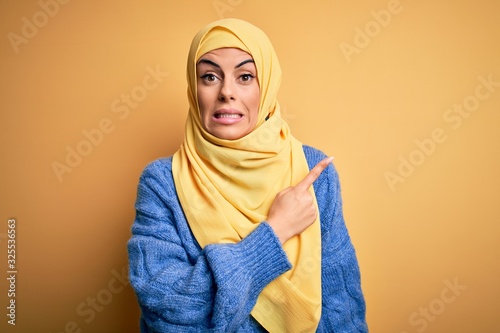 Young beautiful brunette muslim woman wearing arab hijab over isolated yellow background Pointing aside worried and nervous with forefinger, concerned and surprised expression © Krakenimages.com