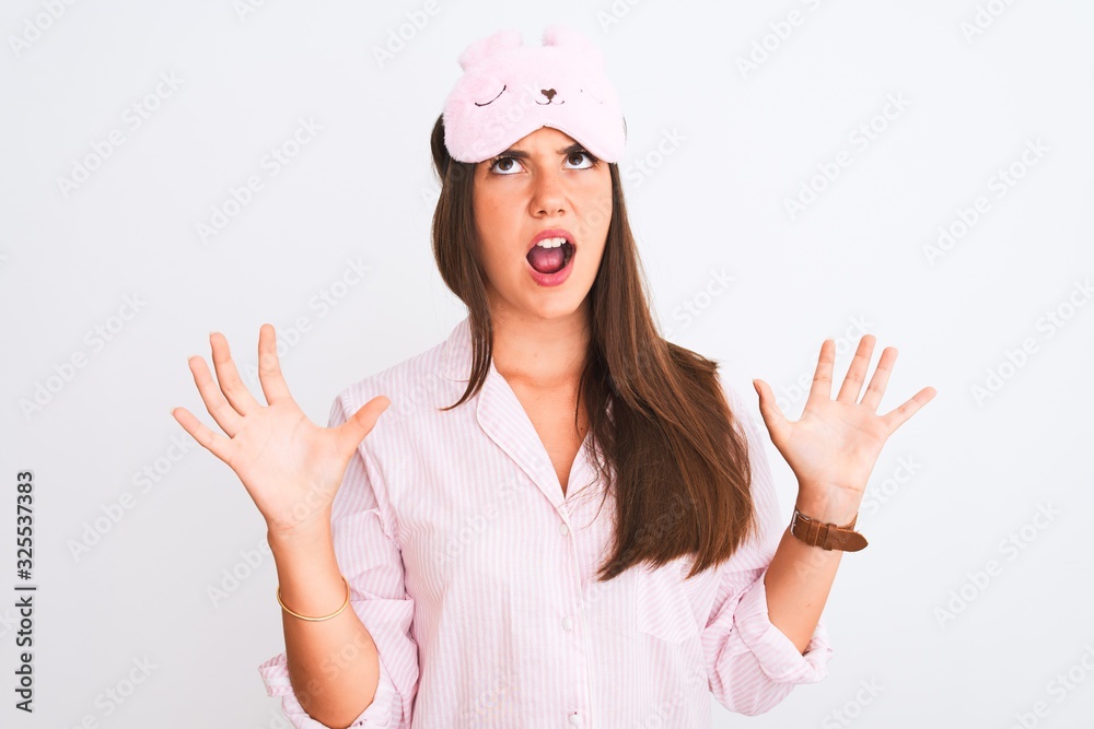 Young beautiful girl wearing pajama and sleep mask standing over isolated white background crazy and mad shouting and yelling with aggressive expression and arms raised. Frustration concept.