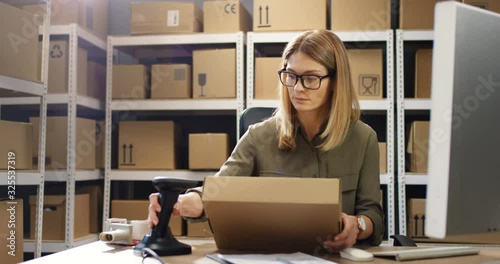 Beautiful Caucasian woman post worker sitting at desk and working at computer in postal delivery store. Female scanning parcel and registering it in system in post office.