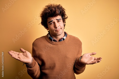 Young handsome man wearing casual shirt and sweater over isolated yellow background clueless and confused with open arms, no idea concept.