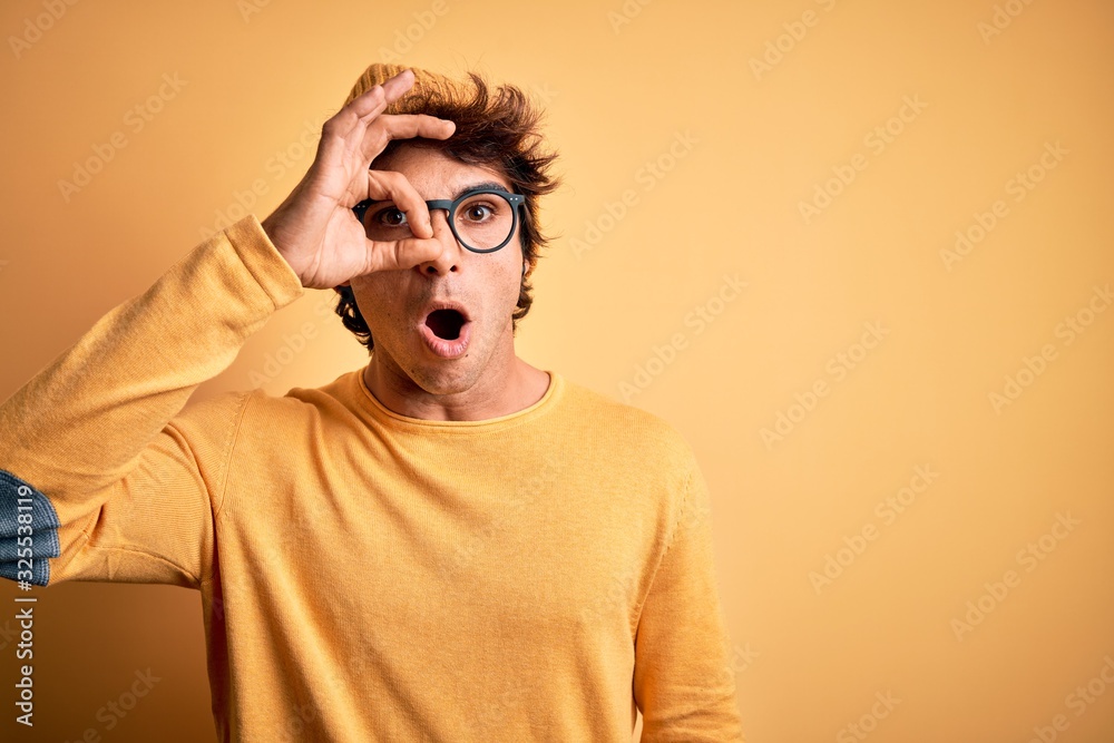 Young handsome man wearing casual t-shirt and glasses over isolated yellow background doing ok gesture shocked with surprised face, eye looking through fingers. Unbelieving expression.