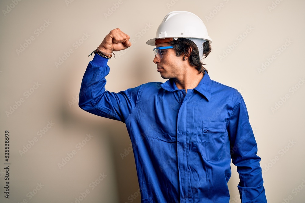 Young constructor man wearing uniform and security helmet over isolated white background showing arms muscles smiling proud. Fitness concept.