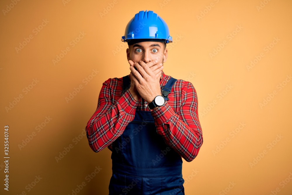 Young builder man wearing construction uniform and safety helmet over yellow background shocked covering mouth with hands for mistake. Secret concept.