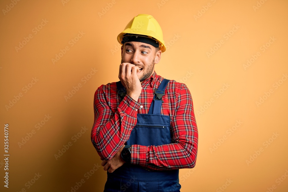 Young builder man wearing construction uniform and safety helmet over yellow isolated background looking stressed and nervous with hands on mouth biting nails. Anxiety problem.