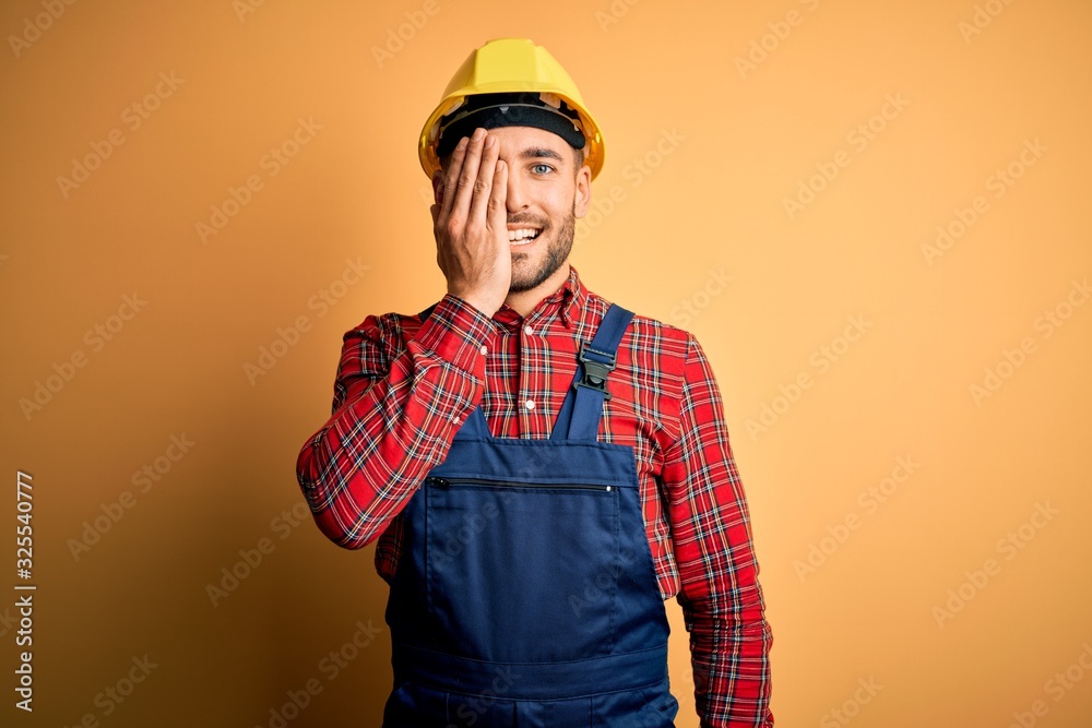 Young builder man wearing construction uniform and safety helmet over yellow isolated background covering one eye with hand, confident smile on face and surprise emotion.