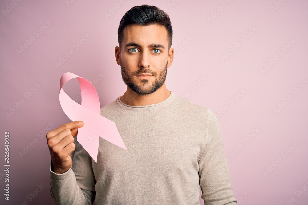 Young handsome man holding pink cancer ribbon symbol of support over isolated background with a confident expression on smart face thinking serious Young handsome man holding pink cancer ribbon symbol of support over isolated background with a confident expression on smart face thinking serious
