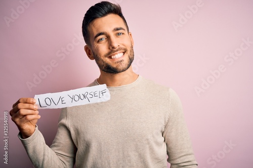 Young handsome man holding paper with self love message over pink background with a happy face standing and smiling with a confident smile showing teeth © Krakenimages.com