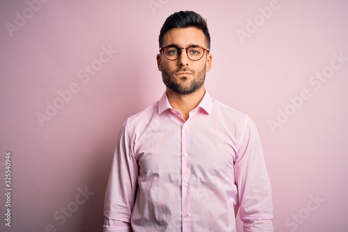 Young handsome man wearing elegant shirt and glasses standing over pink background with serious expression on face. Simple and natural looking at the camera. © Krakenimages.com