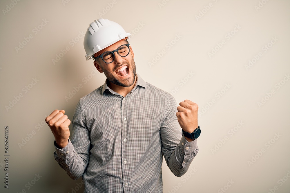 Young architect man wearing builder safety helmet over isolated background very happy and excited doing winner gesture with arms raised, smiling and screaming for success. Celebration concept.