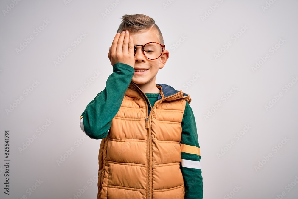 Young little caucasian kid with blue eyes wearing winter coat and smart glasses covering one eye with hand, confident smile on face and surprise emotion. Young little caucasian kid with blue eyes wearing winter coat and smart glasses covering one eye with hand, confident smile on face and surprise emotion.