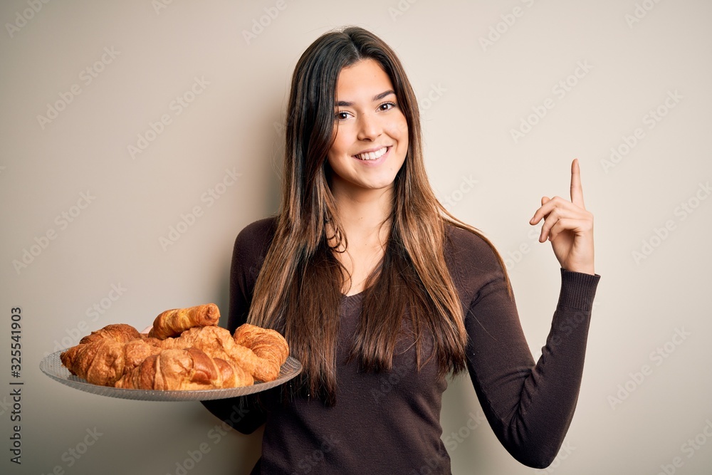 Young beautiful girl holding plate with sweet croissants for breakfast over white background surprised with an idea or question pointing finger with happy face, number one Young beautiful girl holding plate with sweet croissants for breakfast over white background surprised with an idea or question pointing finger with happy face, number one