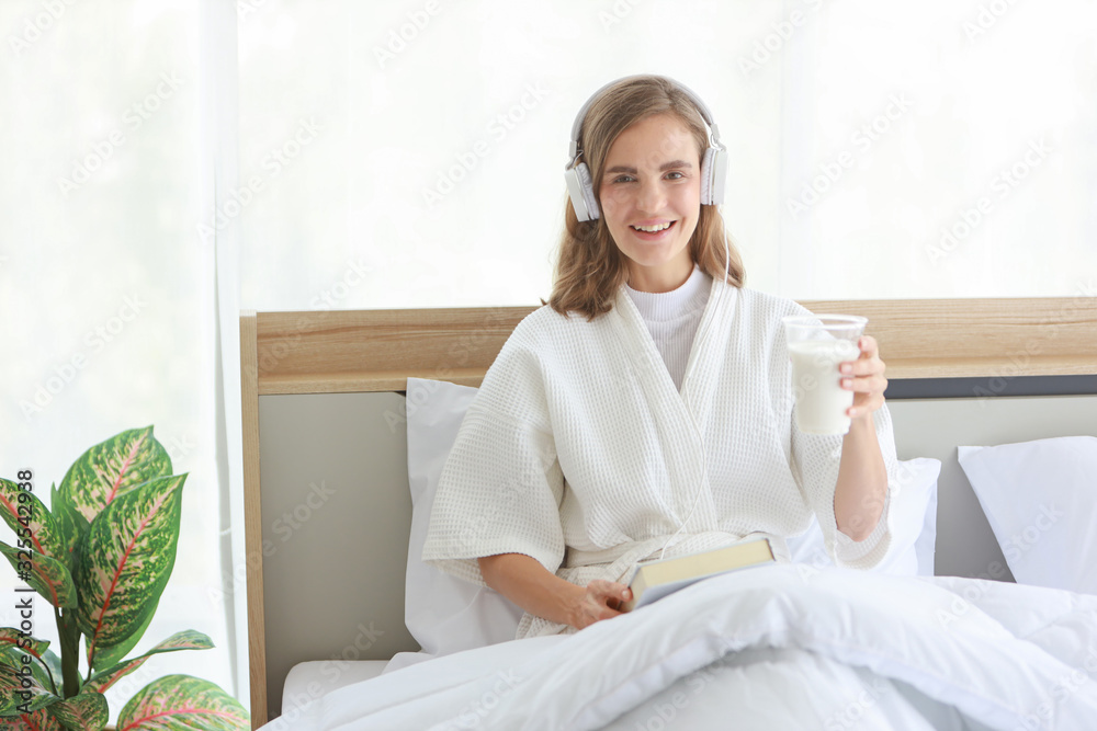 Young cheerful caucasian woman in white pajama with headphone who holding a glass of milk while reading a book before go to sleep with happy and smile face on bed in white modern bedroom. Young cheerful caucasian woman in white pajama with headphone who holding a glass of milk while reading a book before go to sleep with happy and smile face on bed in white modern bedroom.