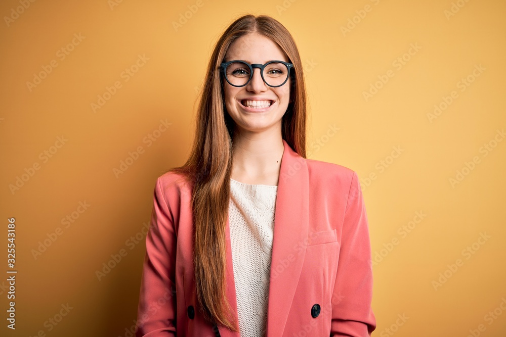 Young beautiful redhead woman wearing jacket and glasses over isolated yellow background with a happy and cool smile on face. Lucky person.